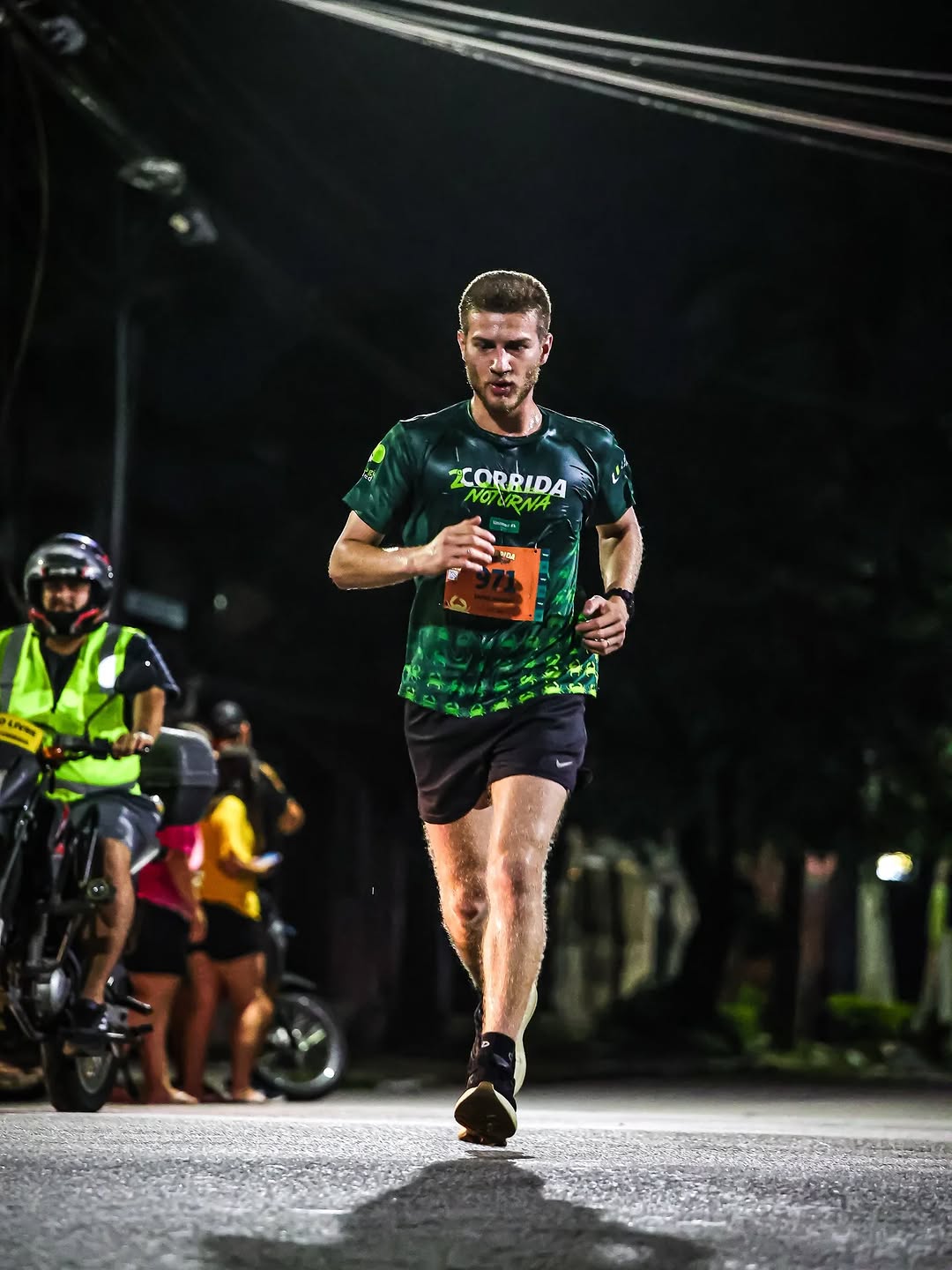 Gabriel correndo em uma rua durante a noite. Ele aparece no centro da foto, em movimento, usando uma camiseta verde de corrida da Unimed, shorts pretos e tênis. No peito, há o número de identificação 971 preso à camiseta. Ele está concentrado, olhando para frente.

Ao fundo, há algumas pessoas acompanhando a corrida, incluindo um motociclista usando capacete e colete refletivo. A rua está iluminada por luzes artificiais, enquanto o restante do ambiente está escuro. A foto foi tirada de um ângulo baixo, próximo ao chão, destacando o corredor em primeiro plano.